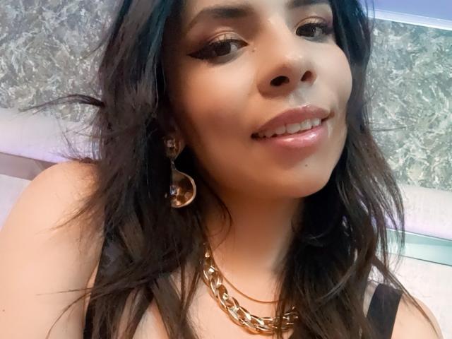 VioletaStone has dark wavy hair, brown eyes, and glossy pink lips. She's wearing gold chain earrings and a chunky gold chain necklace, smiling with a cute nose.