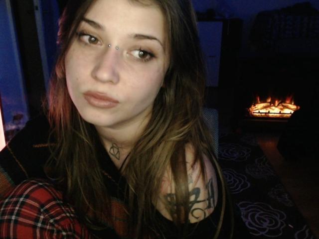 A close-up photo of Alienfortwenty. She has long brown hair, a nose piercing, and a tattoo on her right shoulder. She wears a red and black plaid shirt. Her skin is light, and her expression is neutral. The background shows a lit fireplace with orange flames. The image has a dark, blue-lit setting. Her eyes are slightly up, and her lips are lightly parted. Her left ear is visible. The photo has a casual, intimate feel.