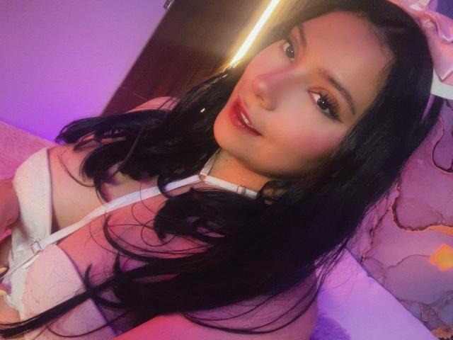 IsabellaCutte with long black hair, wearing a white lace bra, showing her tits, has pinkish-purple lighting on her face, and a subtle smile. Her eyes are slightly open.