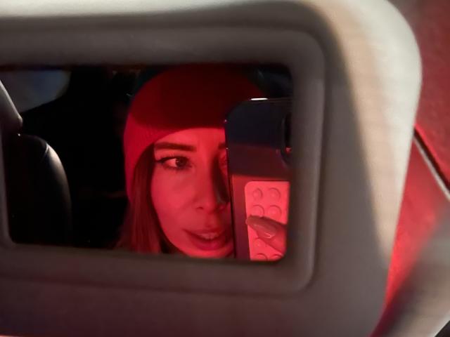 JennyLawry with red beanie, holding phone in car mirror, red lighting, visible tits, slightly open mouth, phone case with heart design, car interior, partial reflection.
