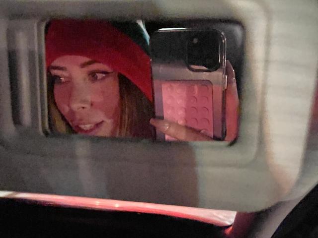 JennyLawry's face in a car mirror selfie, wearing a red beanie, holding a pink phone with a patterned case, looking slightly to the side with a subtle smile.