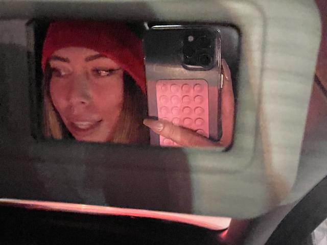 JennyLawry in a red beanie, taking a selfie in a car with a pink-patterned phone, smiling with her finger on the screen. Her tits are partially visible.