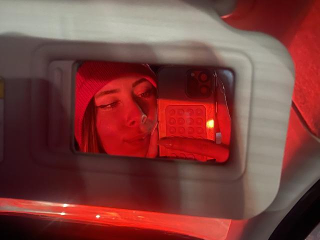 Il viso di JennyLawry riflesso nello specchietto retrovisore dell'auto, indossa un berretto rosso, con un telefono che copre parte del suo viso, una luce rossa che si riflette sulla sua pelle e un leggero sbuffo..