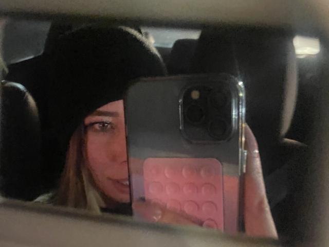 JennyLawry che si fa un selfie in macchina, indossa un berretto nero, una custodia rosa per il telefono con motivi a esagono, si vede un occhio e un sorriso leggero, un iPhone con tre obiettivi per la fotocamera..
