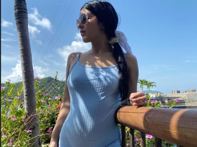AdelleVelvet with dark pigtails, wearing a light blue ribbed tank top, showing her small tits, standing by a wooden railing, wearing sunglasses, in a sunny outdoor setting.