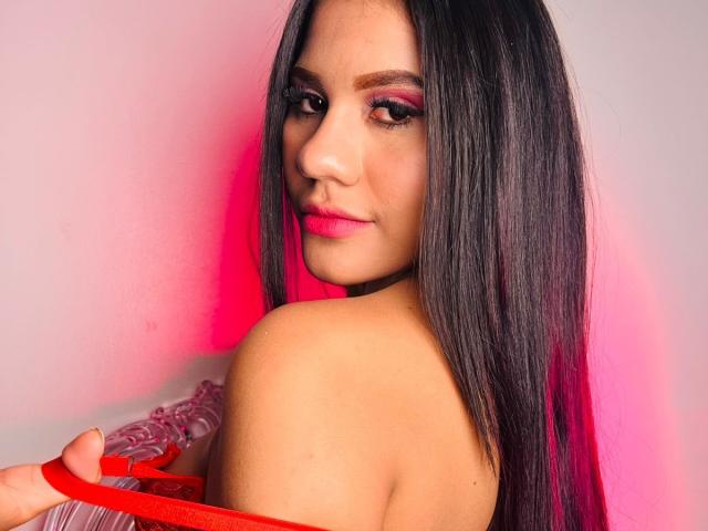 AmeliaSexx with long black hair, pink eyeshadow, red lipstick, and red bra straps, holding bra, looking over shoulder, smiling, pink-red light on her face.