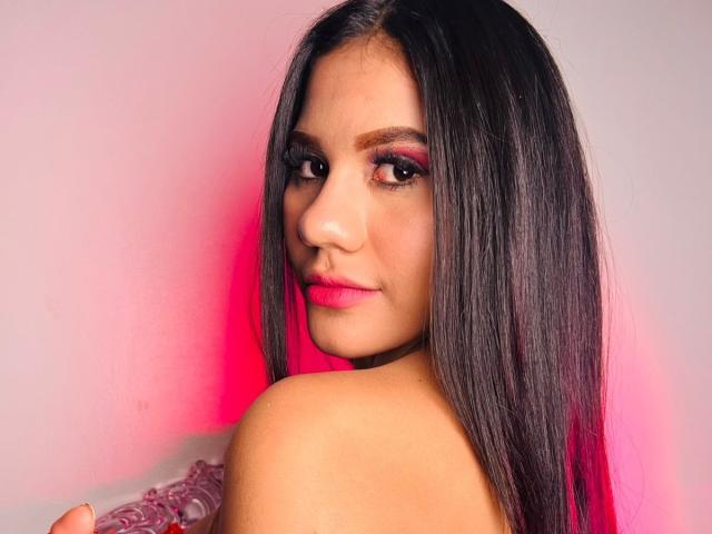 AmeliaSexx with long black hair, pink eyeshadow, and pink lipstick, looking over her shoulder, showing her bare shoulder and a subtle smile, in a well-lit, pink-lit photo.