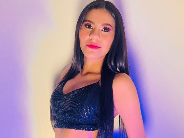 AmeliaSexx with long black hair, wearing a sparkly black crop top, has pink eyeshadow and red lipstick, looking at the camera with a slight smile, showing her tits.