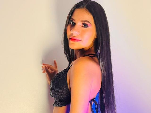 AmeliaSexx with long black hair, red lipstick, and a sparkly black bra, standing against a wall, looking over her shoulder with a slight smile.
