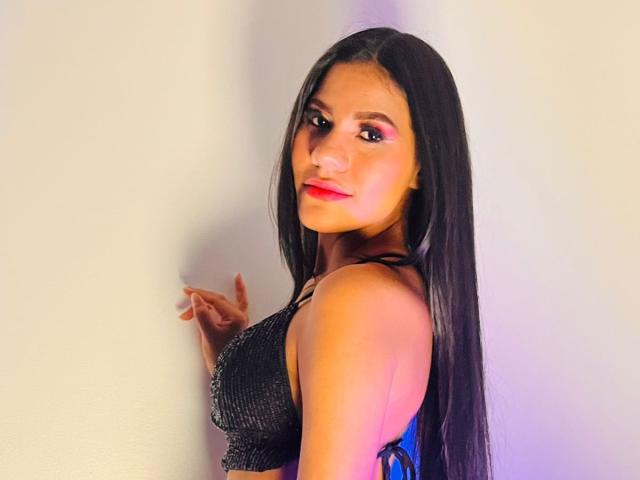 AmeliaSexx with long black hair, wearing a black sequined top, showing her tits, standing against a white wall, smiling with pink lipstick, and looking over her shoulder.