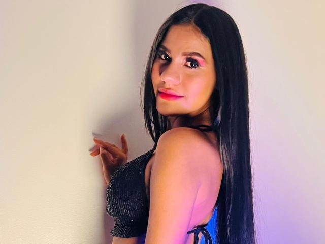 AmeliaSexx with long black hair, red lipstick, and sparkly black top, looking over shoulder, showing her small tits and sideboob, standing against a white wall.