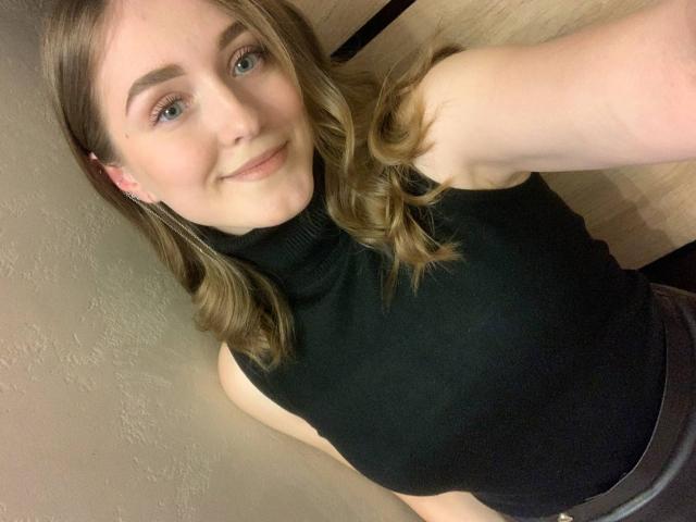 MoonBloom, met blond golvend haar, draagt een zwart mouwloos turtleneck shirt, glimlacht naar de camera en laat haar blote armen en een subtiele blik op haar achterwerk zien..