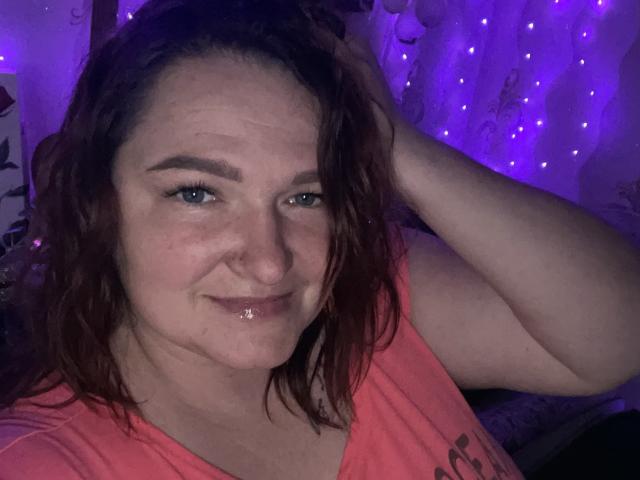 AngelQSky has wet brown hair, blue eyes, and a slight smile with shiny lips. She's wearing an orange shirt, her hand in her hair, under purple lights.