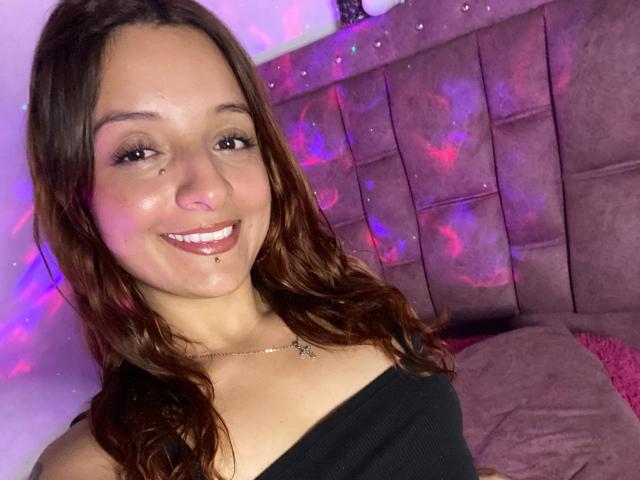 SaarahJones with wavy brown hair, smiling, has a nose piercing and is wearing a black off-shoulder top, showing her tits. She has a silver necklace and pinkish-purple lighting.
