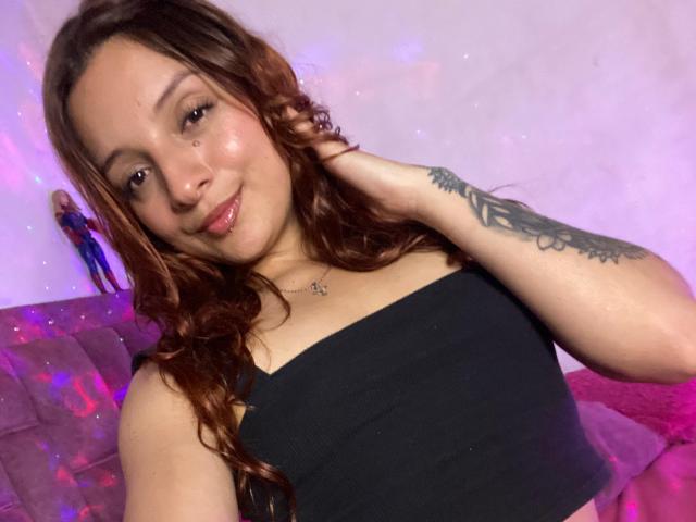SaarahJones with curly brown hair, black tube top, tattoo on right arm, smiling, pinkish-purple lighting, holding hair, pink couch in background, small nose piercing, cute face.