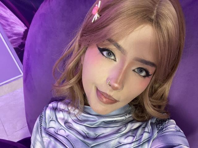 Carla Miceli tem cabelos castanho-rosados, olhos azuis com delineador grosso, um piercing no nariz e está vestindo uma blusa prateada, de estilo futurista, com uma textura metálica brilhante..