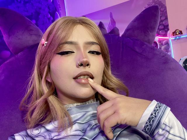 CarlaMiceli with pink hair, black winged eyeliner, nose ring, and pink hairclip, biting her finger, wearing a sparkly choker and blue patterned shirt, purple background.