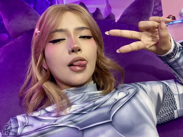 Carla Miceli, con i capelli biondi, un piercing al naso e rossetto rosa, con la lingua fuori, indossa un'armatura argentata e fa un gesto di pace, seduta su un divano viola, in una foto sexy in stile cosplay..
