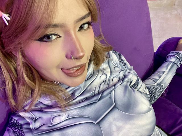 CarlaMiceli with pink hair, nose ring, and tongue out, wearing shiny silver armor, sitting on a purple couch, showing her tits. Her makeup is bold with black eyeliner.