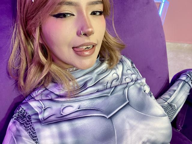 CarlaMiceli with blonde hair, nose ring, and winged eyeliner, wearing shiny silver armor, tongue slightly out, purple chair in background, looking cute and confident, showing her tits.