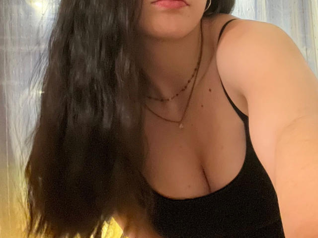 Nyrah-hot with long black hair, wearing a black tank top, showing her large tits and cleavage. She's got a necklace on and is leaning forward slightly.