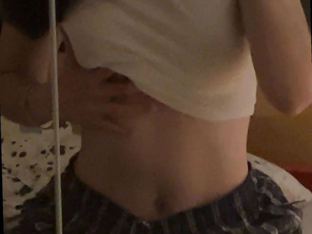 Nyrah-hot's shirt is lifted, exposing her flat, pale belly with a visible navel. She's wearing striped pajama bottoms, her hand slightly covering her right side.