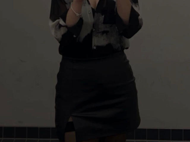 Nyrah-hot with dark skirt, black blouse, and gray scarf, standing, hands raised, visible arms, slightly visible breasts, no pants, tile floor, dim lighting, partial upper body.