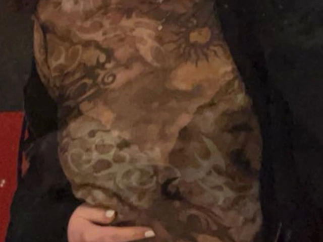 Nyrah-hot's arm with a detailed brown and orange tattoo featuring a sun, animal faces, and swirling patterns. A hand with white-painted nails is holding the arm.