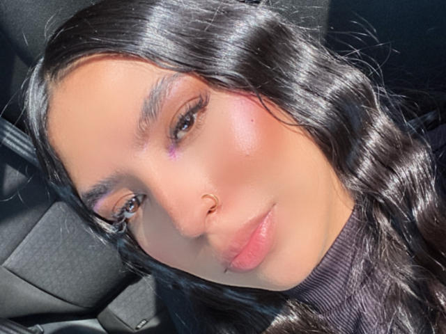 O rosto de DannaeGray, com cabelo preto brilhante, blush rosado, sombra roxa nos olhos, cílios grossos, lábios rosados, um piercing no nariz e um leve sorriso, olhando diretamente para a câmera..