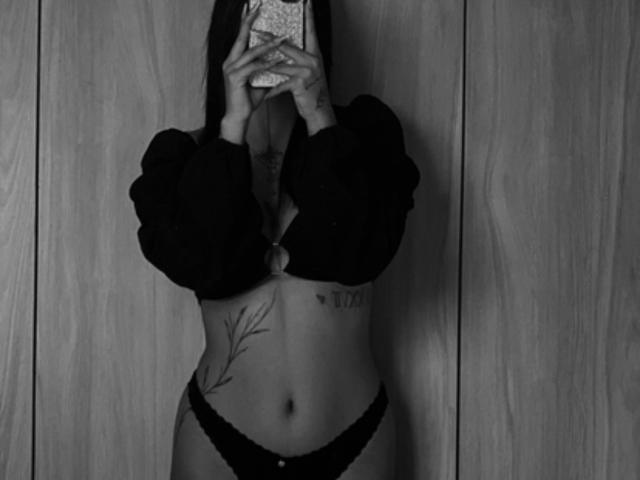 Foto em preto e branco de DannaeGray com mangas bufantes, calcinha de renda e abdômen tatuado, segurando um telefone em frente ao rosto, mostrando seus seios e vulva de forma discreta..