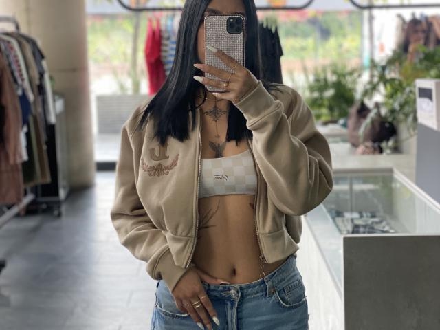 DannaeGray, con i capelli neri, indossa una giacca bomber color cuoio, un top corto bianco che mostra il seno e jeans blu, e sta scattando un selfie allo specchio in un negozio, tenendo in mano un telefono scintillante..