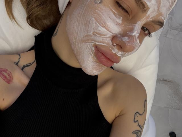SensualMila with white facial mask, winking, black sleeveless top, tattoos on arms, and pink lipstick tattoo. She's lying on a white pillow, looking cute.