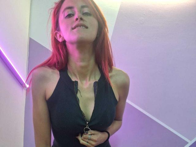 IvanaNovoa with red hair, black zip-up top, showing small tits, slight smile, purple and pink lighting, holding top zipper, wearing rings, looking down at camera.