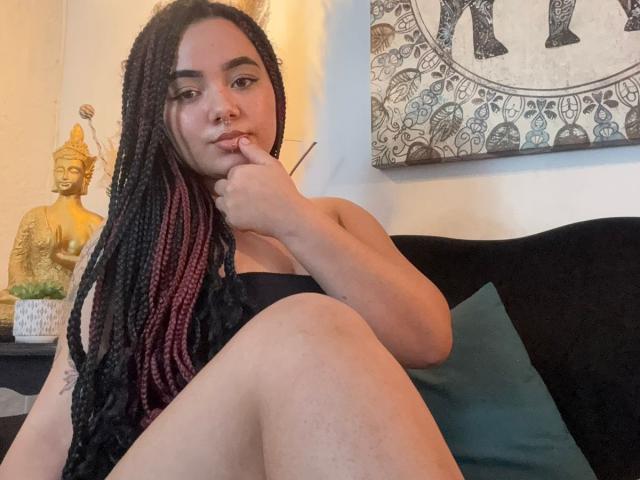 ExoticLuna with black and red braids, nose ring, and finger in mouth, sitting with legs up, wearing black underwear, on a black couch with teal pillow.
