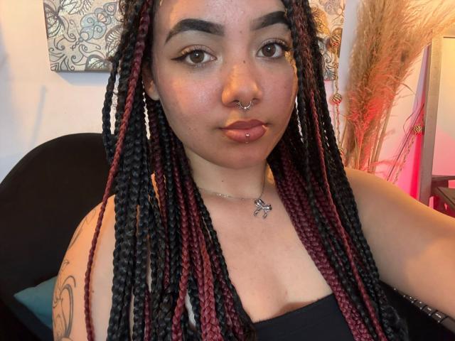 ExoticLuna has dark braids with red and black sections, a nose ring, and a lip ring. She's wearing a black top with a silver necklace, and has a tattoo.