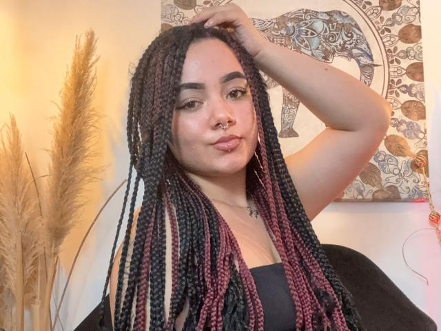 ExoticLuna has long braided black and pink hair, a nose ring, and is pouting with her arm raised. She's wearing a black top, showing her tits slightly.