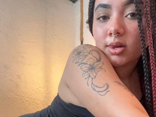 ExoticLuna with braided hair, nose ring, lip piercing, and tattoo on shoulder, wearing black top, looking at camera, slightly open mouth. Her skin is light brown, she's attractive.