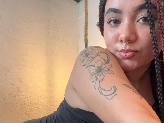 ExoticLuna with braided hair, nose ring, lip piercing, and tattoo of flowers on her shoulder, wearing a black top, looking at the camera with a slight pout.