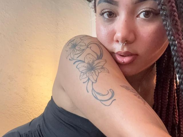 ExoticLuna with braided hair, nose ring, and glossy lips, showing off a floral tattoo on her shoulder. She's wearing a black top, her arm's raised.
