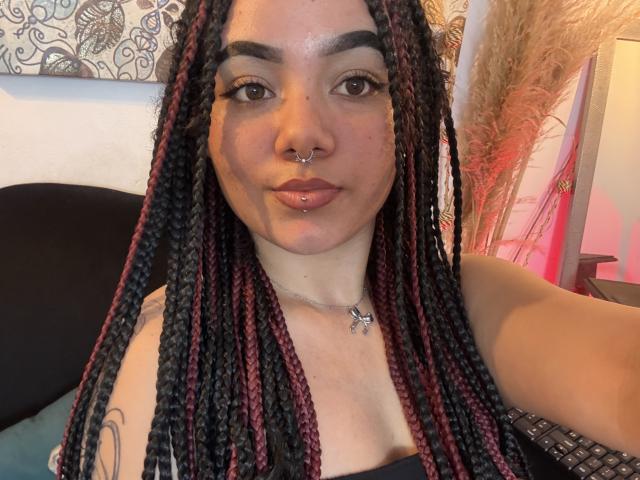 ExoticLuna has dark braided hair with red highlights, a nose ring, and a lip piercing. She's wearing a black top, showing her tits and a tattoo on her arm.