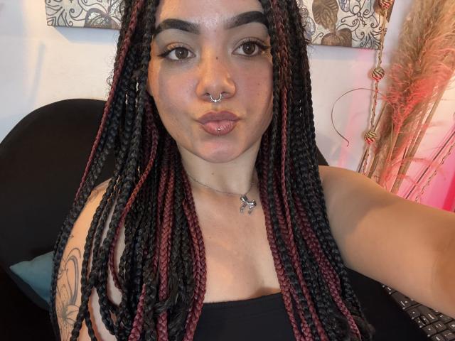 ExoticLuna with black and red braided hair, nose ring, and lip piercing, wearing a black top, showing a tattoo on her arm, pouting her lips.