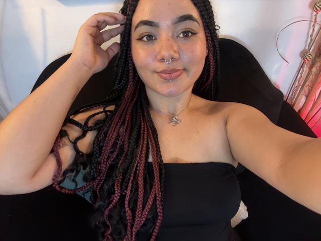 ExoticLuna with dark braided hair, red and black tips, nose ring, and lip piercing, wearing a black strapless top, smiling, hand in hair, black chair, pink light.