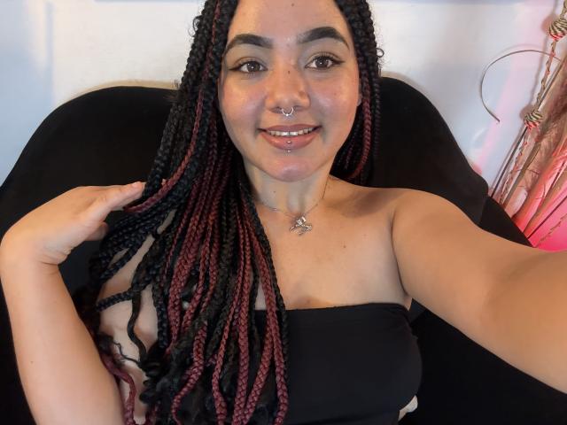 ExoticLuna with braided black and red hair, nose ring, and lip ring, smiling, holding hair, wearing black strapless top, sitting in black chair, necklace with star pendant.