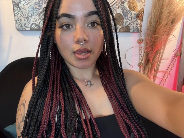 ExoticLuna with braided black and red hair, nose piercing, and lip piercing, wearing a black top, showing a tattoo on her arm, and a necklace with a small pendant.