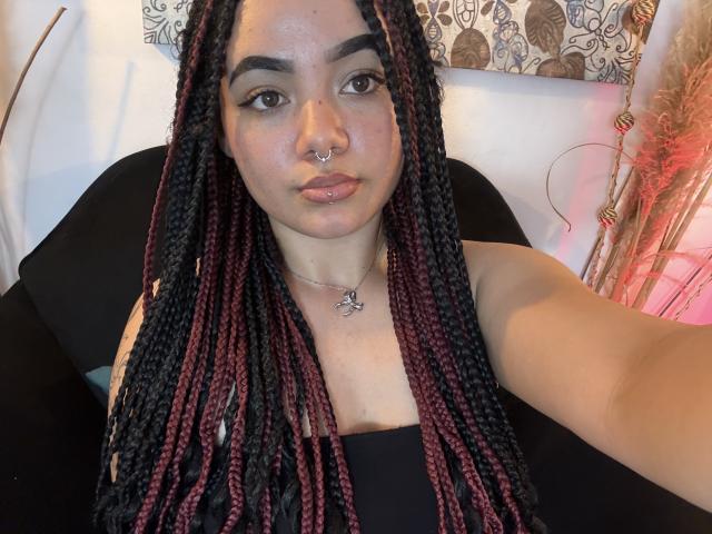 ExoticLuna with black and red braided hair, nose piercing, and lip ring, wearing a black top, showing her tits and a butterfly necklace, taking a selfie.