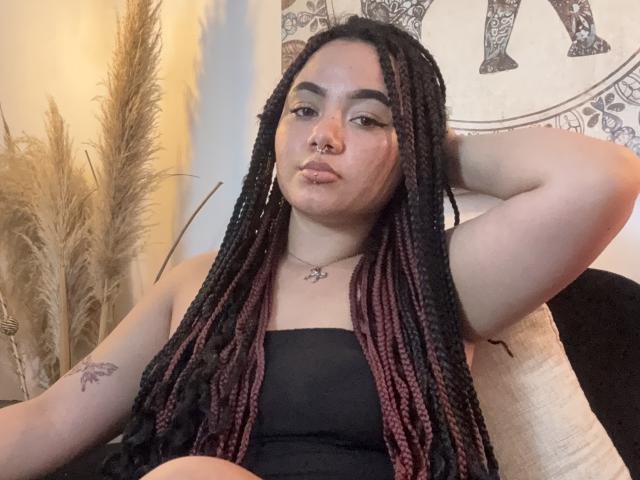 ExoticLuna with long black and pink braided hair, nose ring, and tattoo on her right arm, sitting with hand behind head, wearing a black strapless top.