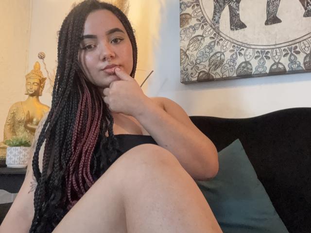 ExoticLuna with braided hair, finger in mouth, black top, sitting on black couch, legs spread, showing bare thigh, gold Buddha statue, and patterned wall art in background.