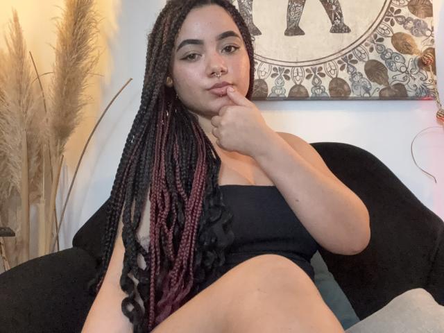 ExoticLuna with long black and red braids, wearing a black tube top, sits on a black couch, finger to her lips, showing her cute nose ring and sexy cleavage.