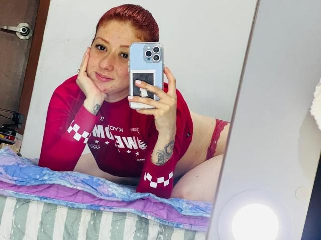 Valeriaaf with red hair, red long-sleeve shirt, and red panties, holding a phone, resting her chin on her hand, sitting on a bed, looking into a mirror.