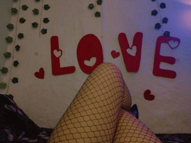 Cami69s' legs in fishnet stockings, knees crossed, showing black high heels, against a wall with red heart decorations.